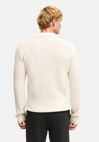 Maglione lavorato a maglia di colore crema con un motivo testurizzato, maniche lunghe e orlo dritto, indossato sopra pantaloni grigio scuro.