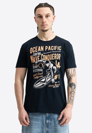 Joven con el cabello trenzado que lleva una camiseta gráfica negra con un surfista y texto sobre el festival de surf Ocean Pacific.