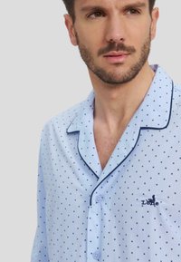 Camisa de pijama de algodón azul claro con lunares azules oscuros, cierre de botones y cuello en contraste. Presenta un logo bordado en el pecho.