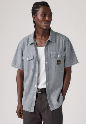 Mand med fletninger iført en kortærmet, stribet skjorte med knapper over en hvid tanktop, stående med den ene hånd i lommen.
