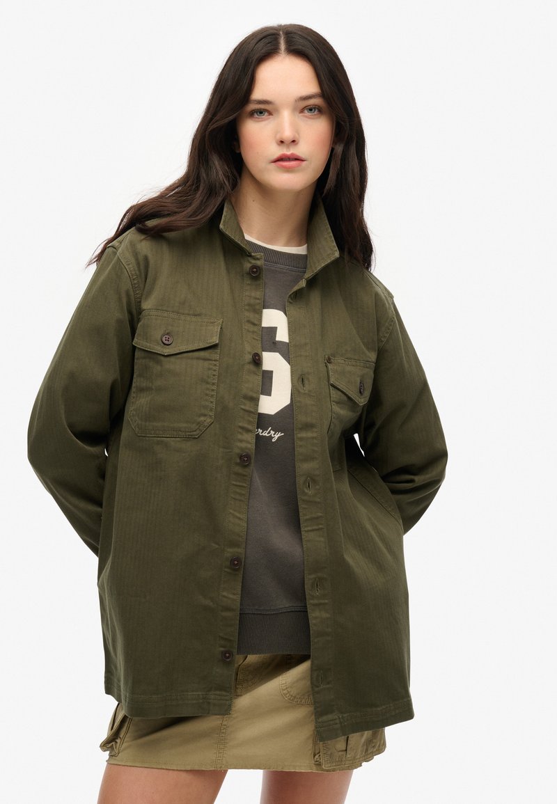 Giovane donna con lunghi capelli scuri, che indossa una giacca verde oliva con bottoni, una felpa scura con stampa del numero e una mini gonna beige.