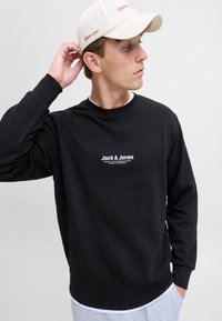 Černá mikina s kulatým výstřihem a dlouhými rukávy, vyznačující se bílým logem "Jack & Jones" uprostřed. Měkká tkanina a volný střih.