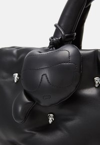 Sac à main en cuir noir avec un visage brodé portant des lunettes de soleil, décoré de petites figurines en argent, et détail de poignée nouée.