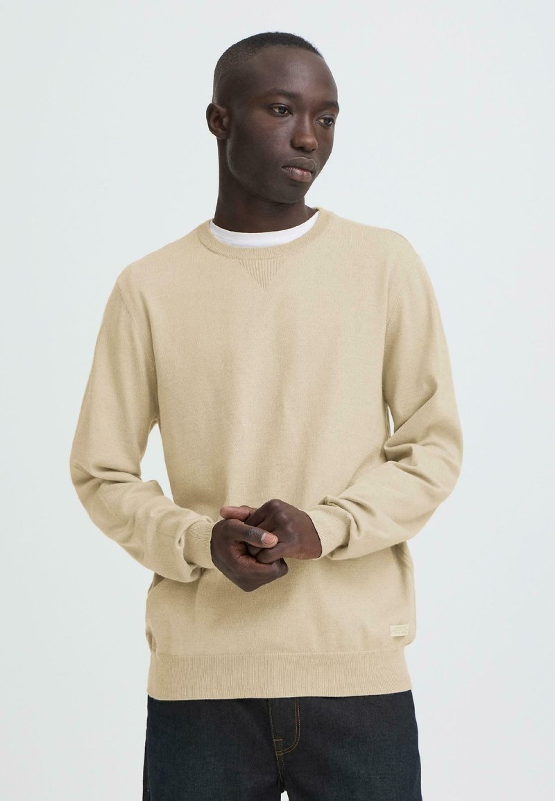 Homme portant un pull beige à col rond et un jean foncé, debout les mains jointes, regardant sur le côté devant un fond uni.