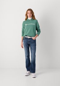 Felpa verde con logo bianco, abbinata a jeans di denim blu e sneakers bianche. La felpa ha una vestibilità comoda e polsini a costine.