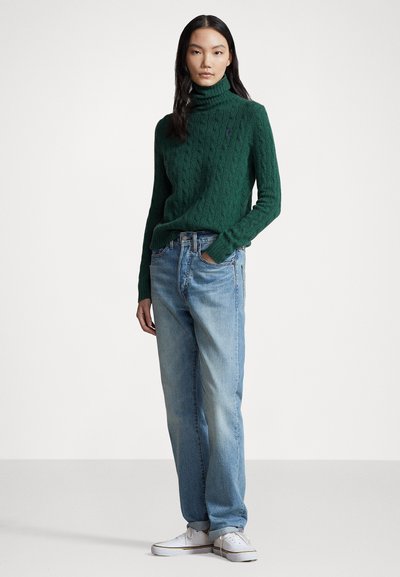 Zöld cable-knit garbó pulóver, amely világoskék farmernadrággal van párosítva. Az öltözet tartalmaz fehér sportcipőt, és lazább szabású.