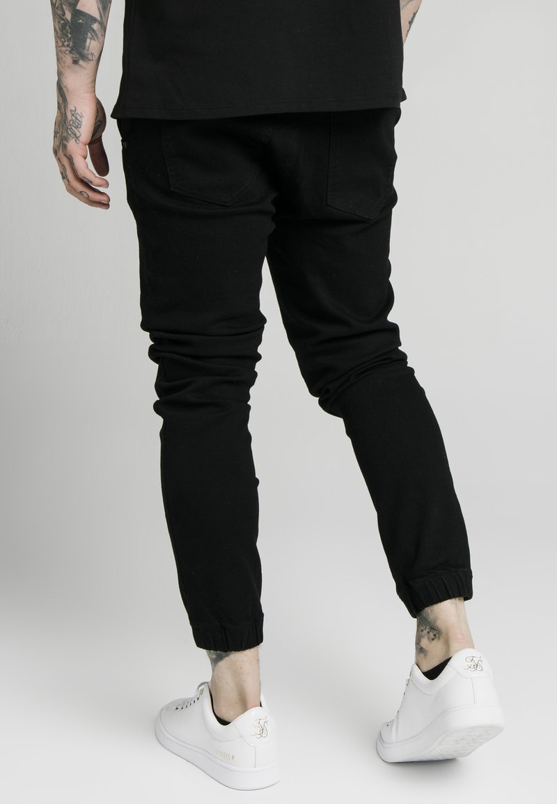Pantalones de chándal negros con un diseño ajustado, puños elásticos y dos bolsillos traseros, combinados con zapatillas blancas con un acento dorado.