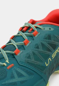 Shoe atlética em teal com um upper texturizado, acentos contrastantes em vermelho e amarelo, e atacadores refletivos. Apresenta uma biqueira reforçada e ventilação em malha.