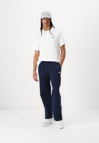 Weißes Baumwoll-T-Shirt mit grünem Logo,navyfarbene Sweatpants mit elastischem Bund und graue Mütze; weiße Turnschuhe runden das Outfit ab.