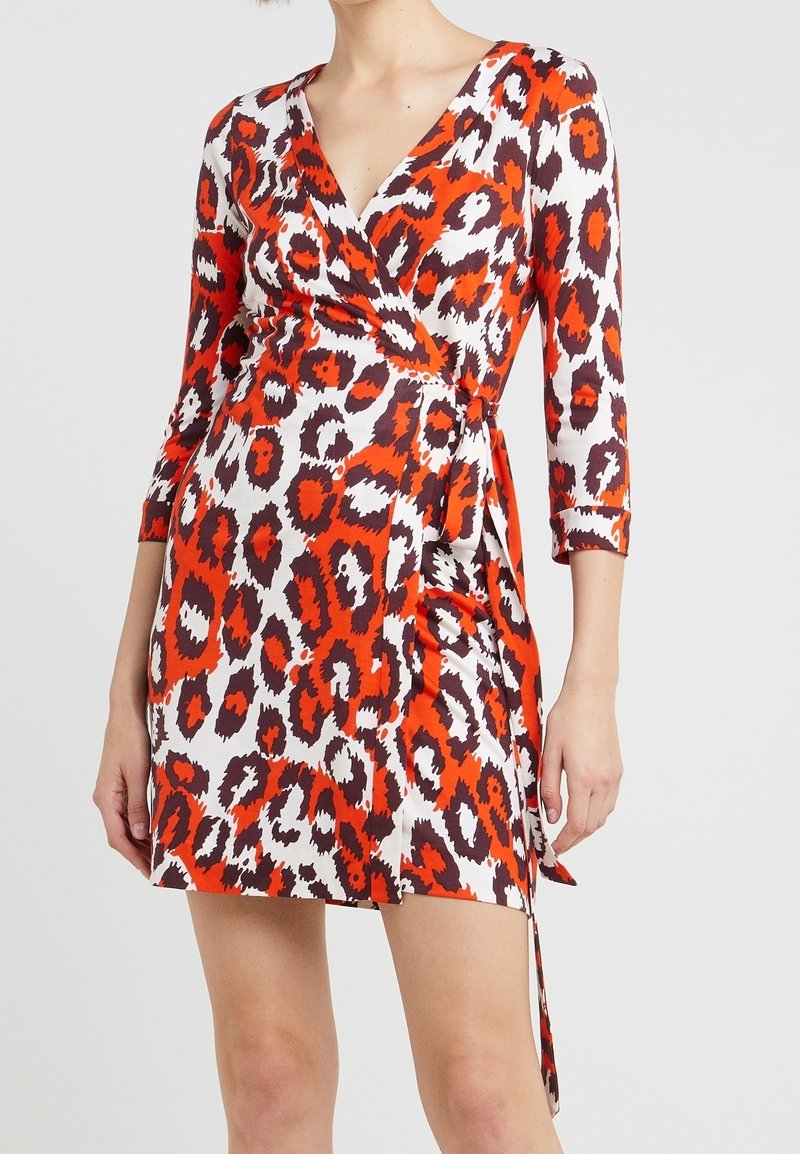 Femme portant une robe portefeuille imprimé animal en rouge, blanc et noir, avec des manches trois-quarts et un décolleté en V, sur un fond clair uni.