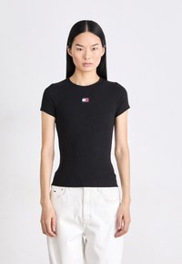 Maglietta nera aderente a maniche corte, con un piccolo logo rosso, bianco e blu sul petto, abbinata a jeans bianchi ampi.
