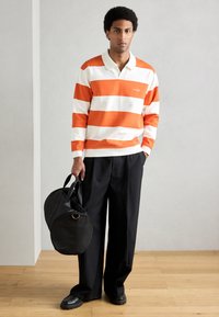 Pólo listrado de manga comprida em laranja e branco, combinado com calças largas pretas. Saco duffel circular preto com duas alças.