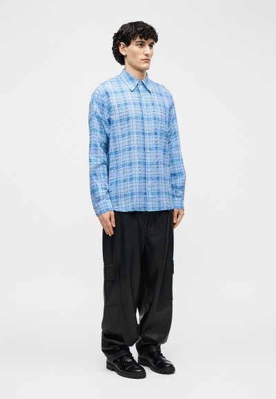 Άνδρας που στέκεται φορώντας μπλε καρό πουκάμισο με μακριά μανίκια, μαύρο φαρδύ παντελόνι cargo και μαύρα παπούτσια, με φόντο λευκό.