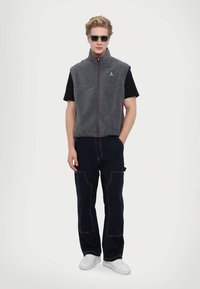 Gray fleece vest with zip, red accents, worn over a black T-shirt. Paired with dark navy pants featuring visible stitching and side pockets.