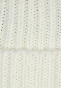 Bonnet tricoté en blanc cassé, présentant un effet de côtes texturé et une finition douce et floue. Le design met en avant des motifs de tricot alternés.