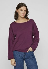 Pull en maille violet avec un large col bateau, des épaules tombantes et des manches longues. Porté avec un jean bleu, doté d'une coupe décontractée et d'une texture.