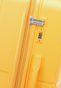 Maleta rígida amarilla con doble cremallera, cerradura de combinación y marca en relieve. Textura suave y forma rectangular.