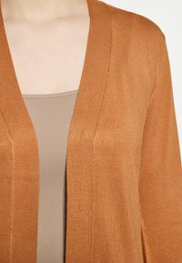 Cardigan de culoare ruginie, cu o textură striată și deschis în față, purtat peste un top de culoare bej deschis.