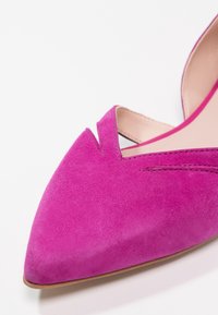 Chaussure plate en suède rose à bout pointu avec un détail en V et doublure intérieure beige sur fond blanc.