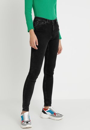 Person trägt eng anliegende schwarze Jeans, grünes geripptes Langarmshirt und weiße Sneaker mit blauen, roten und schwarzen Akzenten, steht vor weißem Hintergrund.