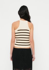 Ribstrikket tanktop i cremefarve med brune vandrette striber. Den har en høj halsudskæring og et åbent rygdesign. Kombineret med et sort blonderet nederdel.
