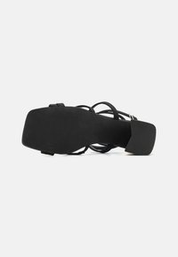 Svart hæl sandal med glatt såle, åpen tå og kryssdesign på stroppene. Teksturet hæl for bedre grep.