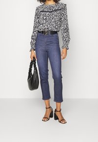 Blusa floreale blu navy con maniche a volant, jeans scuri in denim a vita alta, cintura nera, sandali con tacco neri e una borsa nera.