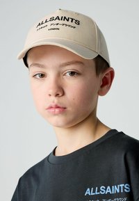 Beige sapka, rajta az "ALLSAINTS" felirattal. Fekete póló logóval, egyszínű és letisztult dizájnnal. Semleges háttér.