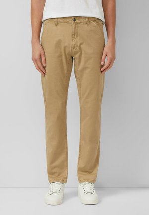 Person wearing tan straight-leg pants, white sneakers, and a white shirt against a plain light gray background.