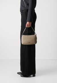 Beige leren handtas met een gladde textuur, ritsclosing en korte handgreep, vastgehouden in een hand naast zwarte wijde broek en hoge laarzen.