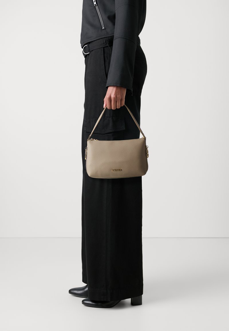 Beige leren handtas met een gladde textuur, ritsclosing en korte handgreep, vastgehouden in een hand naast zwarte wijde broek en hoge laarzen.