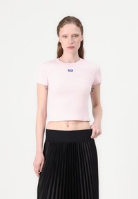Svetloružový rebrovaný crop top s okrúhlym výstrihom a krátkymi rukávmi, s modrou nášivkou loga na hrudi. Nosený s čiernou skladanou sukňou.