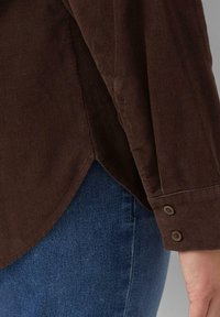 Bruine corduroy blouse met ribbeltextuur en knoopmanchetten, voorzien van een gebogen zoom en een iets relaxte pasvorm. Gecombineerd met blauwe denim.