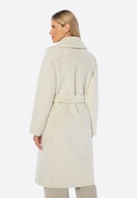 Abrigo largo color crema con una superficie lisa y texturizada, cuello chal y cinturón en la cintura, con un corte de longitud media.