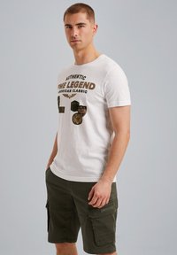 T-shirt en coton blanc avec texte imprimé et graphiques de badge ; associé à un short cargo vert foncé avec poches latérales et détails de couture.