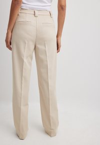 Beige maßgeschneiderte Hose mit strukturierter Webart, geradem Beinschnitt und zwei Gesäßtaschen. Der Bund hat Gürtelschlaufen zur Einstellung.