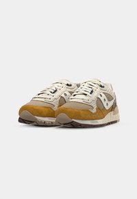 Beige and tan athletic shoes with suede and mesh material, featuring lace-up design, a rubber sole, and contrasting darker brown accents.