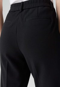 Gros plan sur une personne portant un pantalon noir ajusté avec passants de ceinture et deux poches passepoilées à l'arrière.