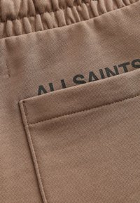 Ruskea puuvillakangas, jossa on teksturoitu pinta, varustettuna takataskulla ja "ALL SAINTS" -painatuksella rohkeilla mustilla kirjaimilla vyötärön läheisyydessä.