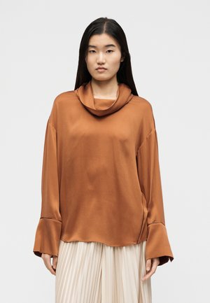 Žena s dlhými tmavými vlasmi, oblečená v voľnej saténovej blúzke hrdzavej farby so širokými manžetami a smotanovej plisovanej sukni, stojaca na neutrálnej pozadí.