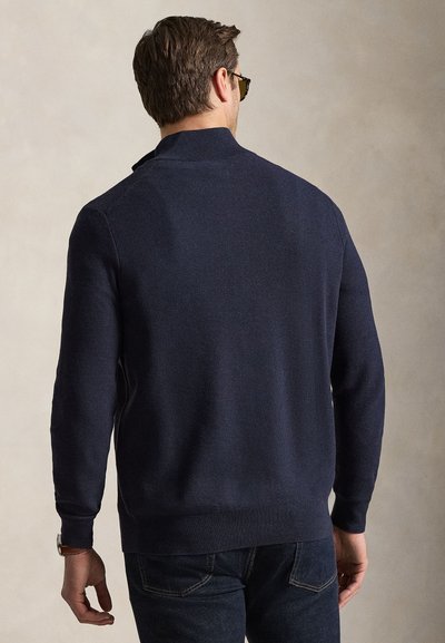 Homme portant un pull à fermeture éclair bleu marine foncé et un jean foncé, vu de dos sur un fond uni beige.