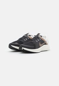Zapatillas deportivas de malla negras con acentos blancos y beige, que presentan un acabado texturizado, punta redonda y cordones en contraste.