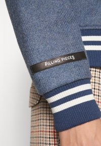 Blå sweatshirtsärm i strukturerad tyg med en svart läderetikett som står "Filling Pieces" och kontrasterande blå och vita randiga ribbade manschetter.