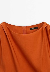 Tissu orange rouille avec encolure bateau et plis près des épaules, avec une étiquette noire avec texte blanc à l'intérieur du col.