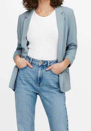 Vrouw met een lichtblauwe blazer, een wit geribd tanksopje en hooggetailleerde lichtblauwe spijkerbroek met handen in de zakken.