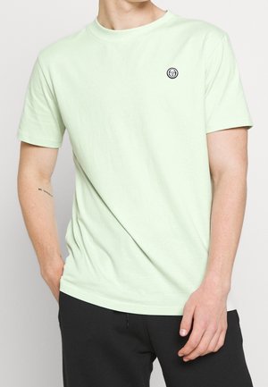 Hellgrünes Baumwoll-T-Shirt mit kurzen Ärmeln und abgerundetem Ausschnitt. Auf der linken Brust befindet sich ein kleines schwarzes rundes Logo.