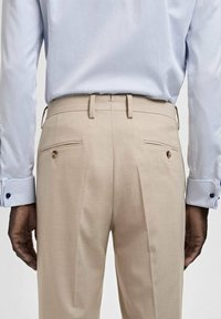 Pantaloni beige su misura con doppie pieghe, due tasche posteriori con bottoni, abbinati a una camicia blu chiaro con colletto. Texture liscia.