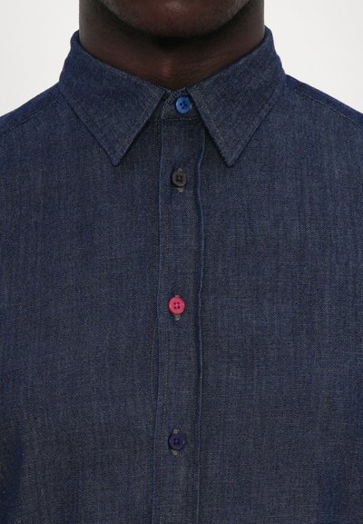 Dark blue denim shirt with a structured collar. Features a row of buttons in light blue, black, pink, and purple. Smooth texture.