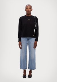 Felpa nera con logo, maniche lunghe e dettagli con bottoni. Jeans a gamba larga blu. Ballerine nere e piccola borsa nera. Sfondo neutro.