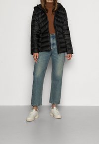 Giacca a vento nera con cappuccio, texture trapuntata, indossata sopra un dolcevita marrone. Abbinata a jeans a gamba dritta di un blu chiaro e sneakers bianche.